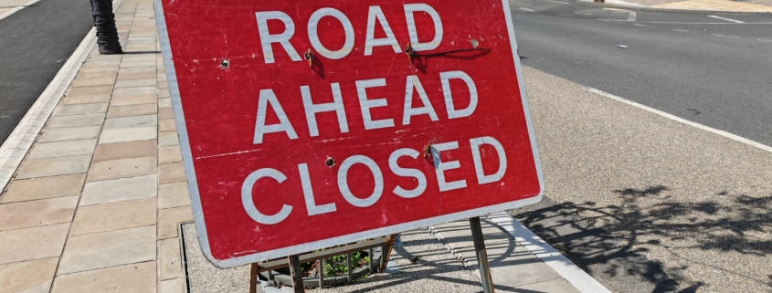 Red sign with "Road Ahead Closed" text stands on a sidewalk. The road nearby is empty, with street furniture and a few people in the background. The scene suggests a temporary road closure in an urban setting.