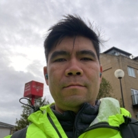 A person wearing a bright neon green jacket and blue earbuds is standing outdoors. The sky is cloudy, and there is a building and trees in the background. They have short dark hair and a serious expression.