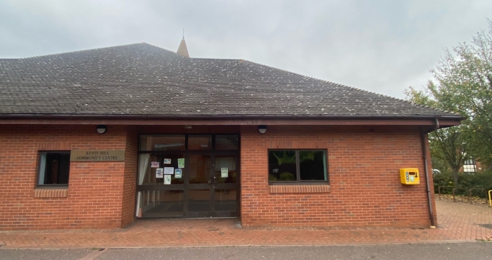 A small brick building with a dark sloped roof labeled "Community Centre" has several notices on the glass door. A yellow defibrillator is mounted on the right side wall. Overcast skies and trees surround the area.