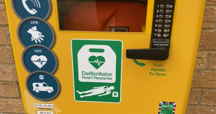 A bright yellow outdoor defibrillator cabinet is mounted on a brick wall. It features instructions for use, including a keypad for an access code, emergency contact number, and icons for calling emergency services, CPR, and using the AED.