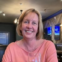 A woman with short blonde hair, wearing a peach top, smiles while sitting in a restaurant or bar, holding a glass. The background includes wooden paneling, dark walls, and television screens.