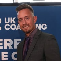 A man in a grey suit and patterned tie smiles in front of a blue background with partial white text that reads "UK COACHING" and "HERE.