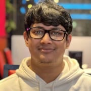 A young man with short dark hair and glasses, wearing a white hoodie, smiles slightly whilst standing indoors with colourful, blurred lights in the background.