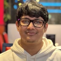 A young man with short dark hair and glasses, wearing a white hoodie, smiles slightly whilst standing indoors with colourful, blurred lights in the background.