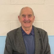 An older man with short grey hair smiles whilst standing against a pale brick wall. He is wearing a grey blazer, a blue jumper, and a checked shirt, and is holding a black folder under his arm.